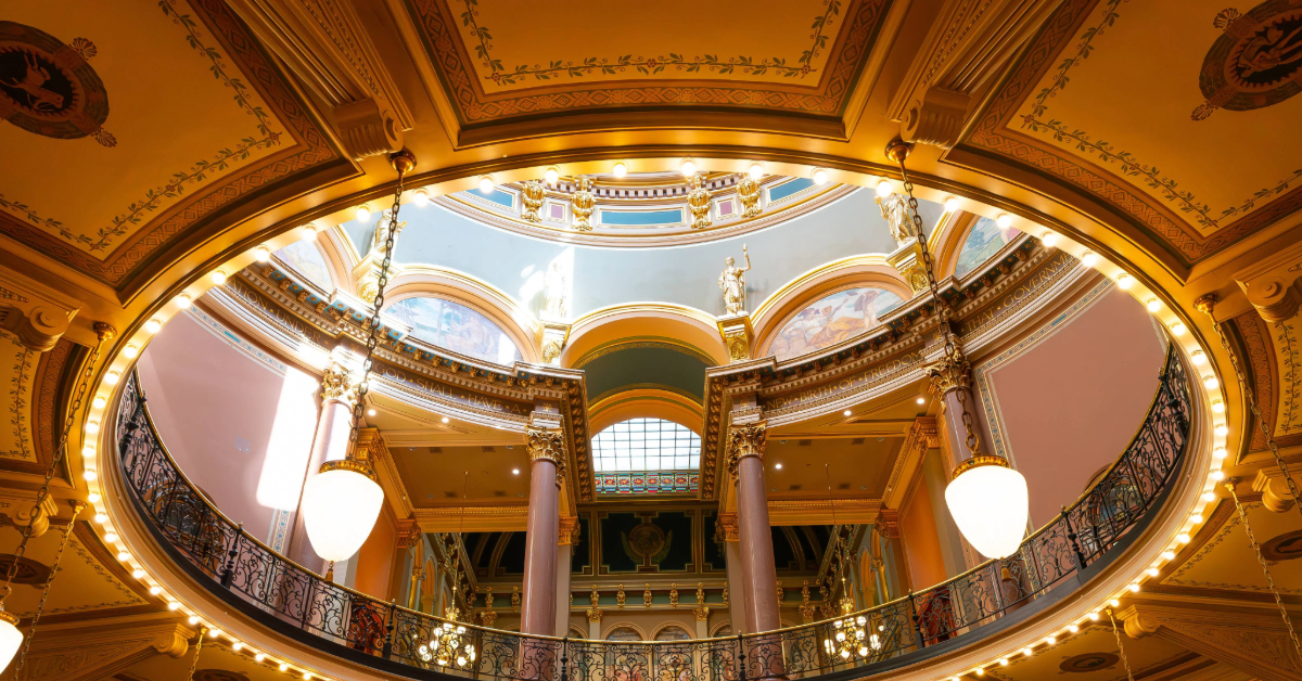 Interior view of an ornate rotunda with decorative arches, columns, chandeliers, and a circular balcony, likely inside a Iowa state capitol building.