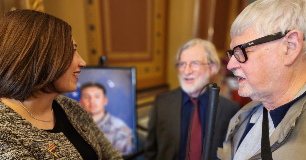 Catelin Drey speaking with Don Wirth inside a formal building, with another person and a blurred portrait visible in the background.